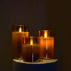 Three amber glass pillar candles are lit on a light blue surface against a dark background.