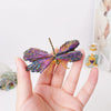 A hand holds a natural crystal tourmaline dragonfly figurine with iridescent purple and blue wings.