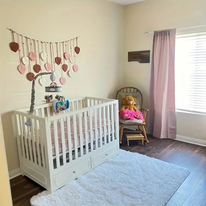 A white crib is positioned next to a wall hanging featuring handwoven pink leaf tassels.