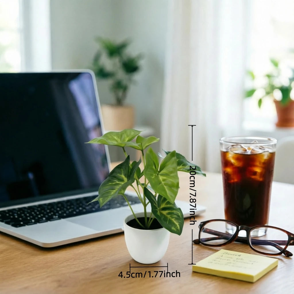 A green artificial potted plant sits on a wooden desk next to a laptop and eyeglasses.