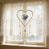A gold heart-shaped pendant with hanging crystal charms is displayed against a sheer white curtain in a window.