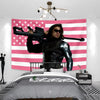 A large pink American flag tapestry wall hanging above a bed, featuring a person in black leather attire holding a rifle.