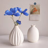 A white ceramic bud vase holds a single blue flower and is displayed next to a white ceramic vase.