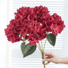 A bouquet of deep red artificial hydrangea silk flowers held by a hand against a white background.