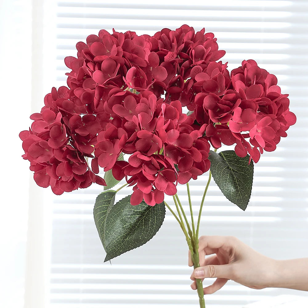 A bouquet of deep red artificial hydrangea silk flowers held by a hand against a white background.