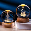 A dark glass crystal ball with a laser engraved scene of a snowy forest and snowmen resting on a wooden base.