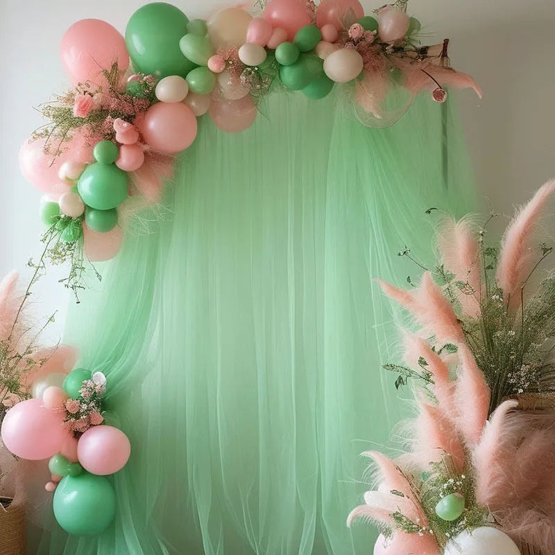 A soft pink and green tulle backdrop decorated with balloons and floral accents for an event.