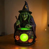 A figurine of a green-skinned witch with long hair holding a staff, sitting behind a glowing green lantern.
