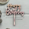 A set of pink and gold acrylic birthday cake toppers spelling out Happy Birthday