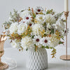 A bouquet of white artificial silk flowers featuring roses, daisies, and greenery displayed in a white vase.
