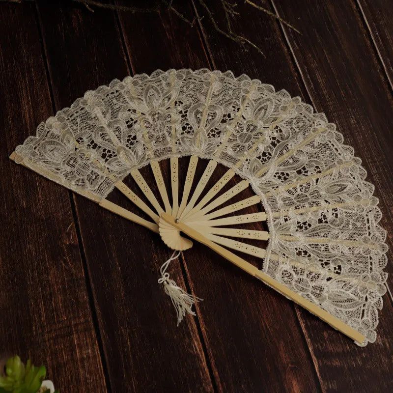 A delicate cream lace folding hand fan with a radiating sunburst design rests on a dark wooden surface.
