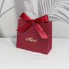 A red paperboard gift bag tied with a satin ribbon and gold script reading Merci, set against a light background.