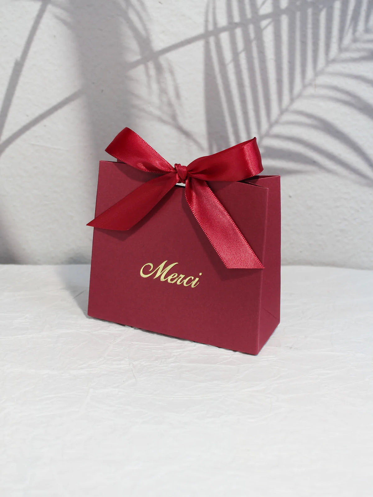 A red paperboard gift bag tied with a satin ribbon and gold script reading Merci, set against a light background.
