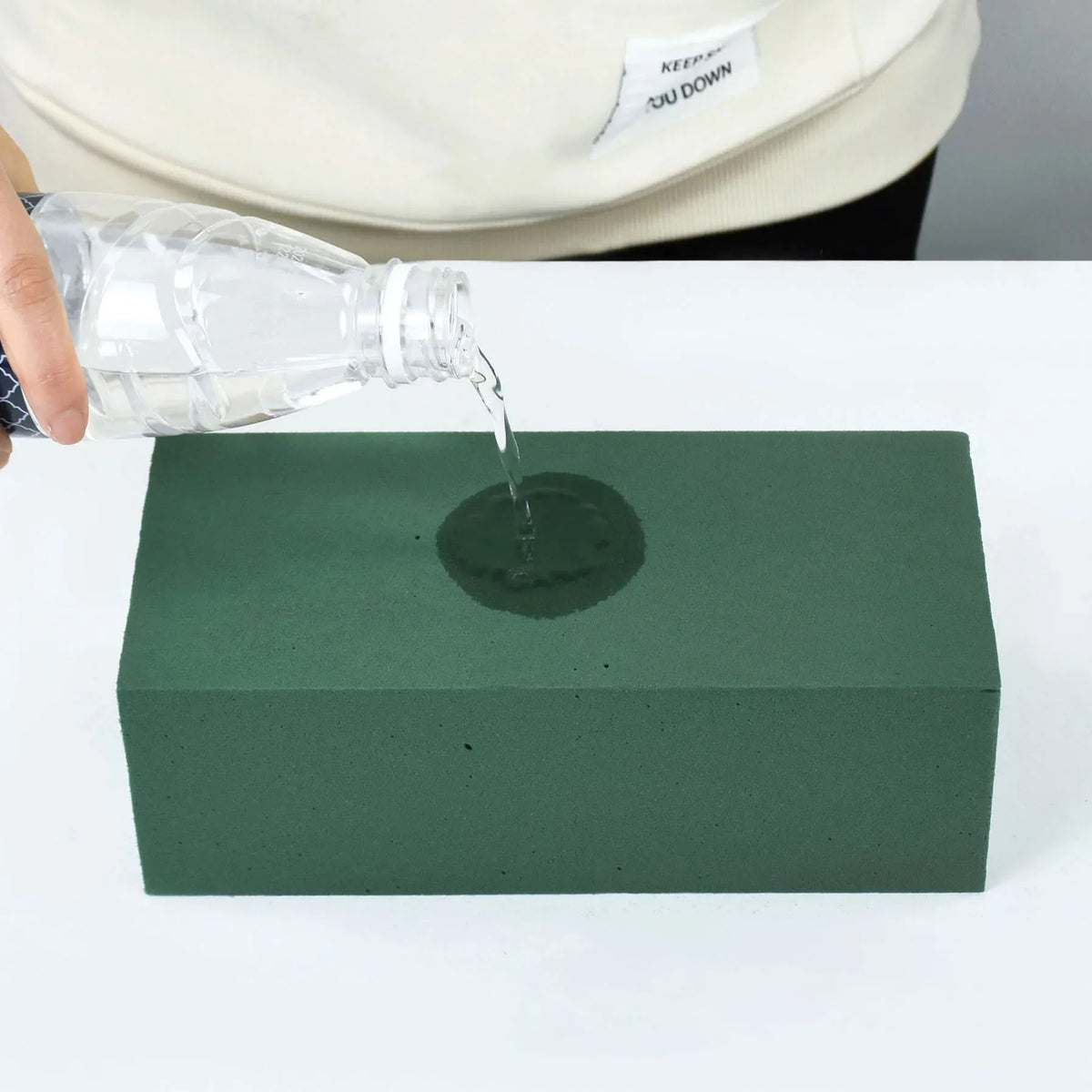 A hand pours water from a plastic bottle onto a dark green foam block for party decor.