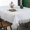 A white embroidered lace tablecloth is set for a meal with floral decorations and tableware on a wooden table.