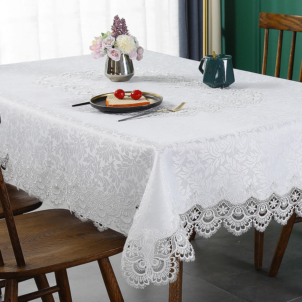 A white embroidered lace tablecloth is set for a meal with floral decorations and tableware on a wooden table.