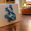 A teal articulated 3D printed frog magnet with orange eyes resting on a wooden surface.