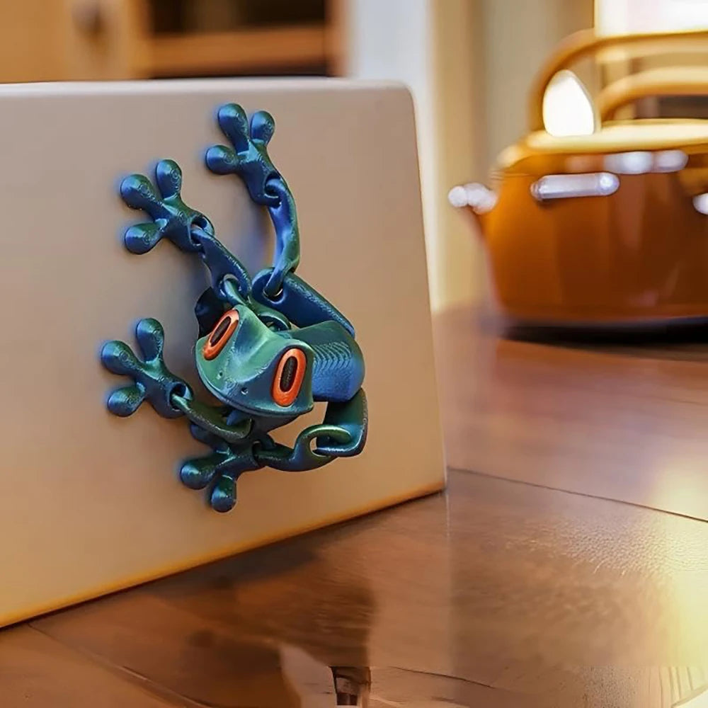 A teal articulated 3D printed frog magnet with orange eyes resting on a wooden surface.
