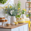 A bouquet of artificial eucalyptus leaf plants with yellow flowers in a white vase on a wooden table.