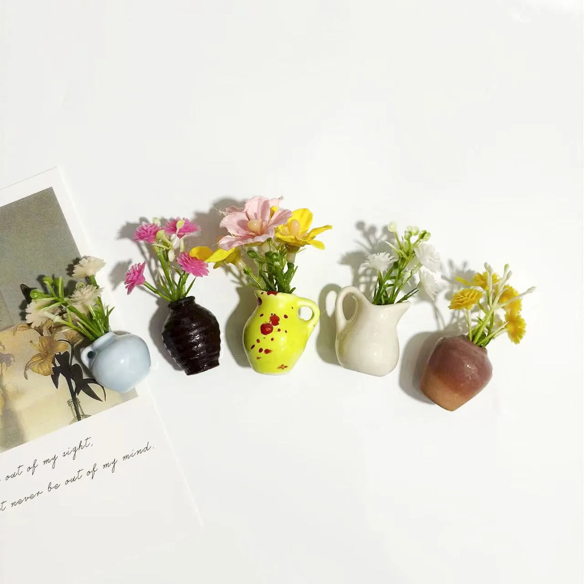 A collection of mini ceramic vases with artificial flowers in yellow, pink, and white on a white background.