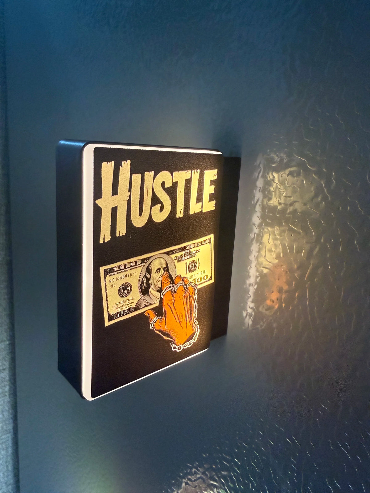 A black sign with the word HUSTLE in distressed white text and a graphic of a hand holding cash, mounted on a wall.