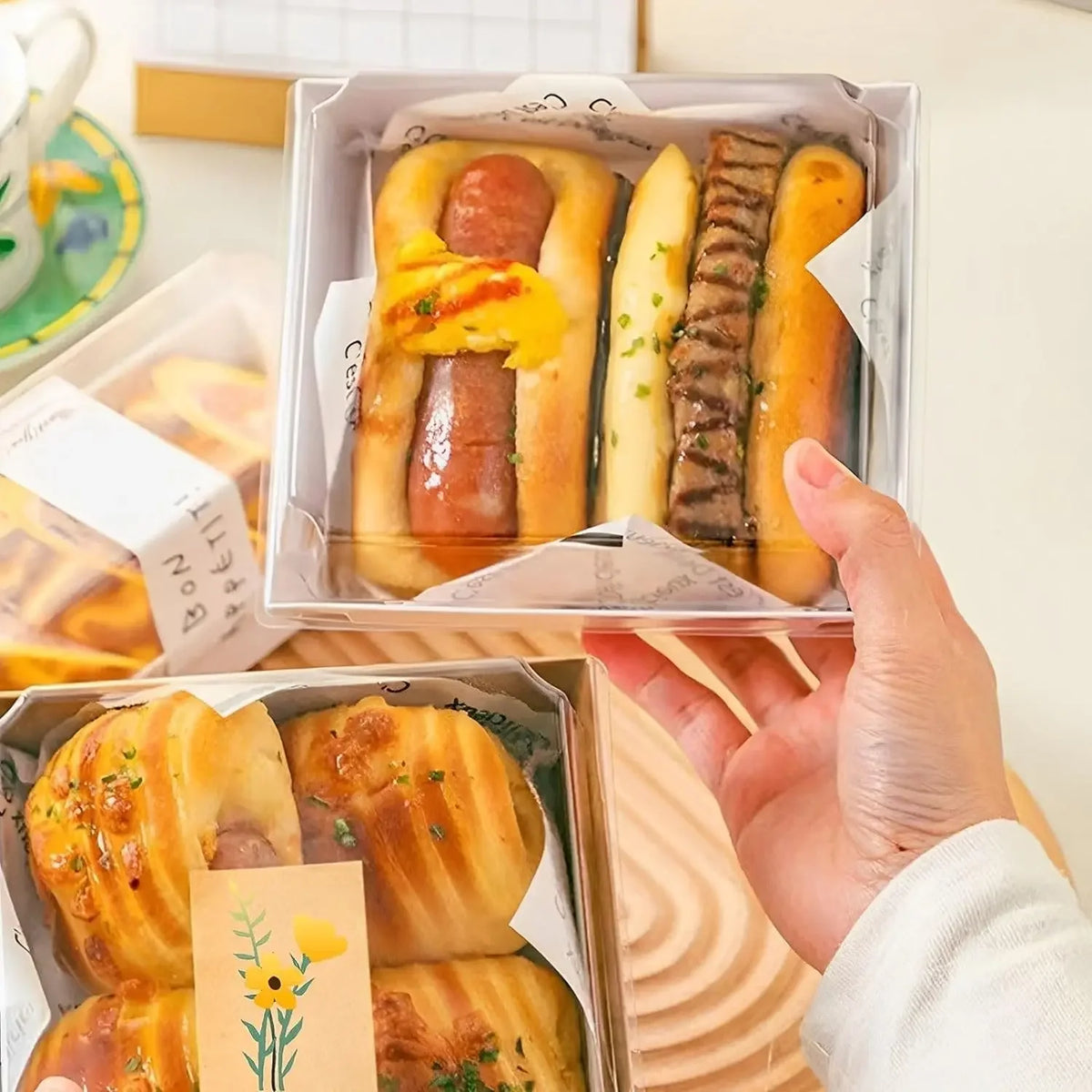 A hand holds an open clear lidded paper sandwich box filled with golden baked pastries and savory items.