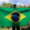 A person holds a large green Brazil flag displaying the national emblem on a sunny outdoor setting.