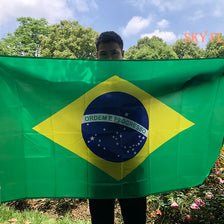 A person holds a large green Brazil flag displaying the national emblem on a sunny outdoor setting.