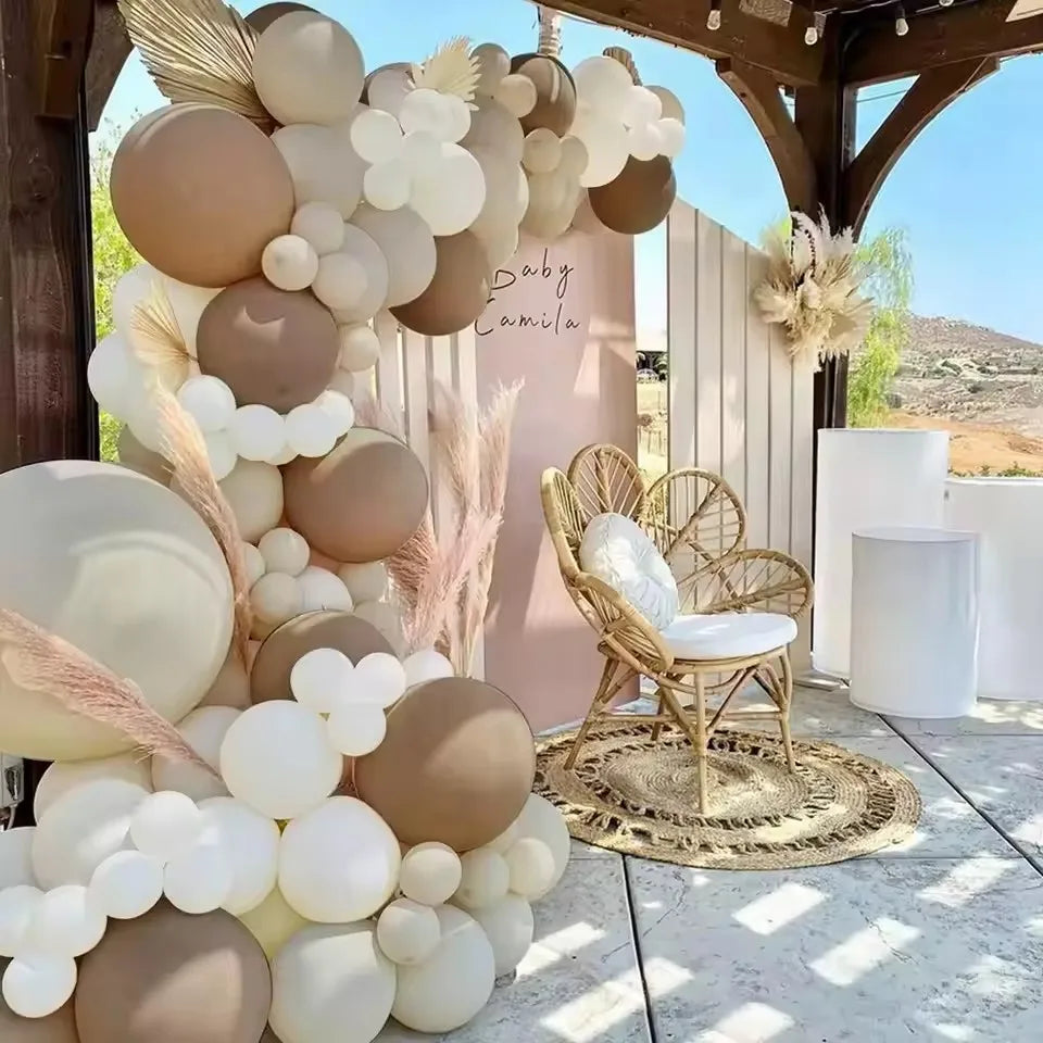 A brown and white balloon garland display in an outdoor setting with a gold chair and wooden archway.