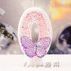A white birthday candle shaped like the number zero decorated with pink glitter and a purple butterfly.