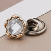 A close-up of a gold-toned brooch button featuring a clear rhinestone set in a floral bezel, resting on a light surface.