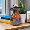 A colorful resin figurine of a frog with large eyes sits on a wooden surface next to a potted plant.