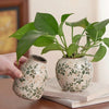 A hand holds a handmade ice crackle ceramic vase filled with green leaves on a light colored surface.