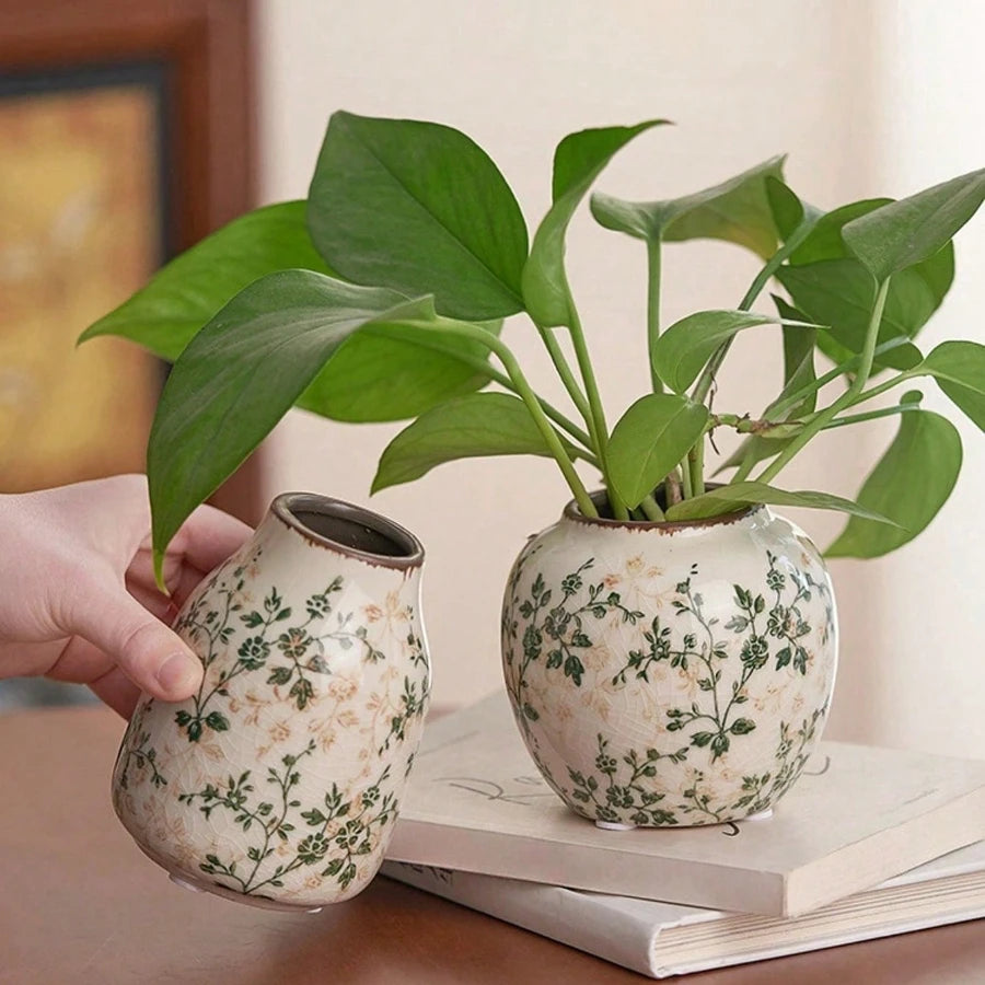 A hand holds a handmade ice crackle ceramic vase filled with green leaves on a light colored surface.