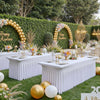 A long white pleated spandex tablecloth cover set up outdoors for an event with greenery and gold decor.