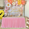 A pink pleated polyester table skirt is displayed on a table, set up for a celebration with balloons and decor.