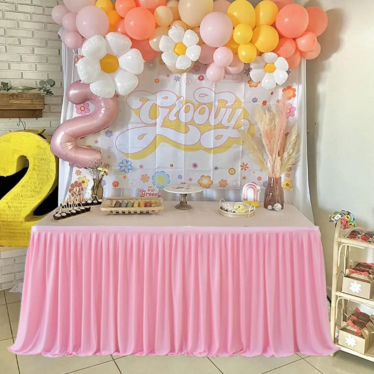 A pink pleated polyester table skirt is displayed on a table, set up for a celebration with balloons and decor.
