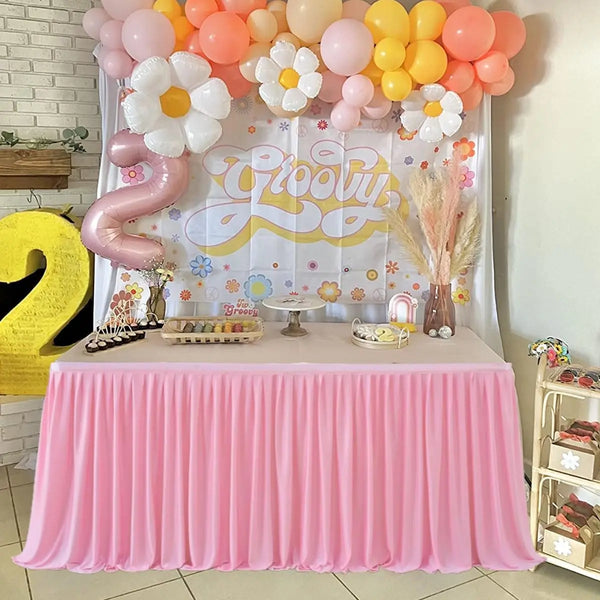 A pink pleated polyester table skirt is displayed on a table, set up for a celebration with balloons and decor.