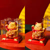 A solar-powered waving lucky cat figurine in a clear box on a red surface.