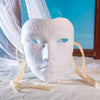 A white masquerade mask with intricate floral designs is displayed on a white cloth.