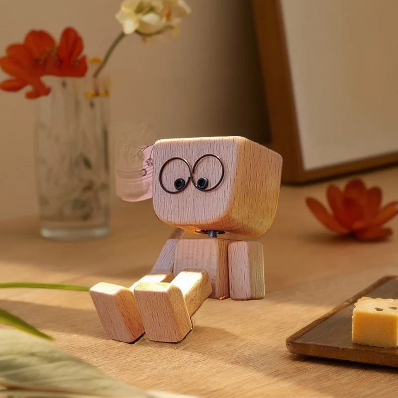 A small wooden figurine of a man with large eyes and pink accents sits on a wooden surface next to flowers.