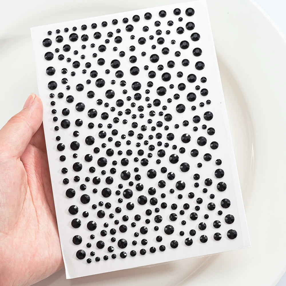 A hand holds a white sheet with black glitter gems arranged in a grid pattern for facial tattoos.