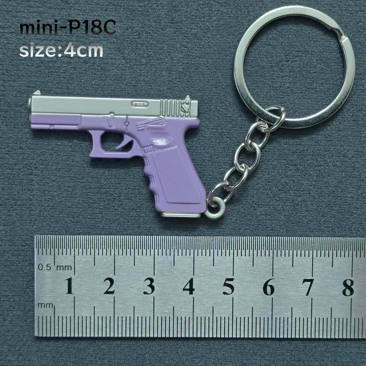 A purple miniature handgun model is attached to a silver keyring, shown next to a ruler.