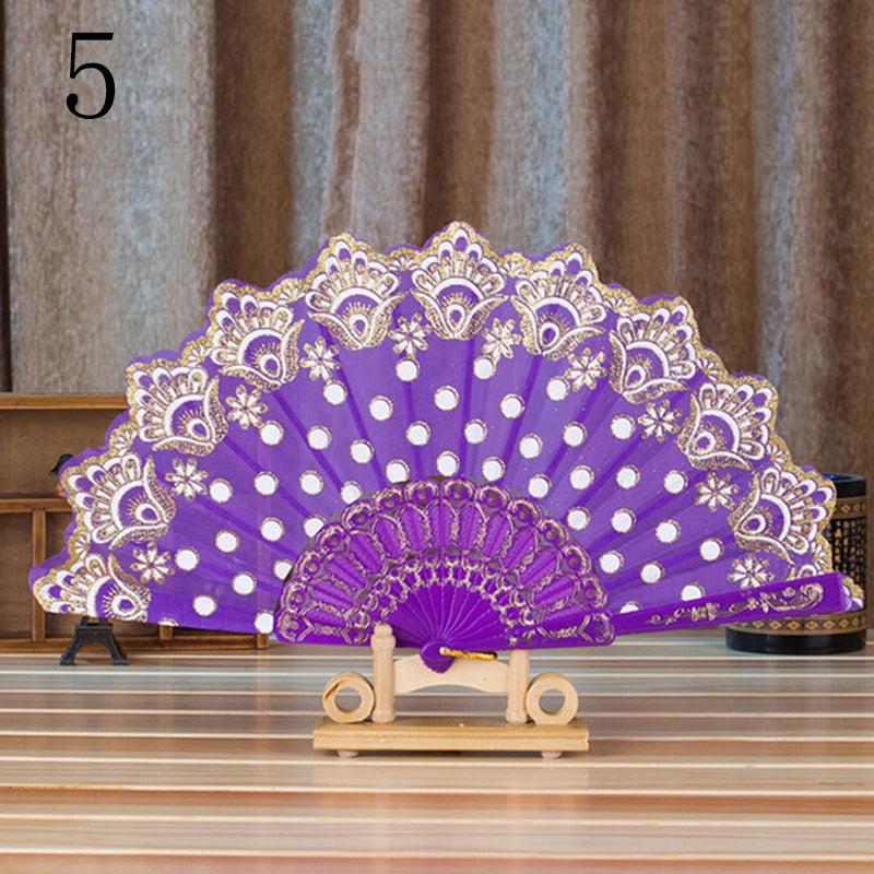 Elegant lace folding hand fan in purple and white with gold detailing displayed on a wooden stand.