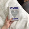 A white photo album titled My World with a heart cutout and the words My Space is held in a hand on a white bed sheet.