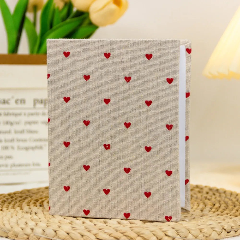 Beige Leinen-Album mit wiederkehrendem Muster aus kleinen roten Herzen, auf einer geflochtenen Matte.