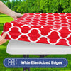 A red patterned waterproof elastic fitted tablecloth is being held over a white table surface outdoors.
