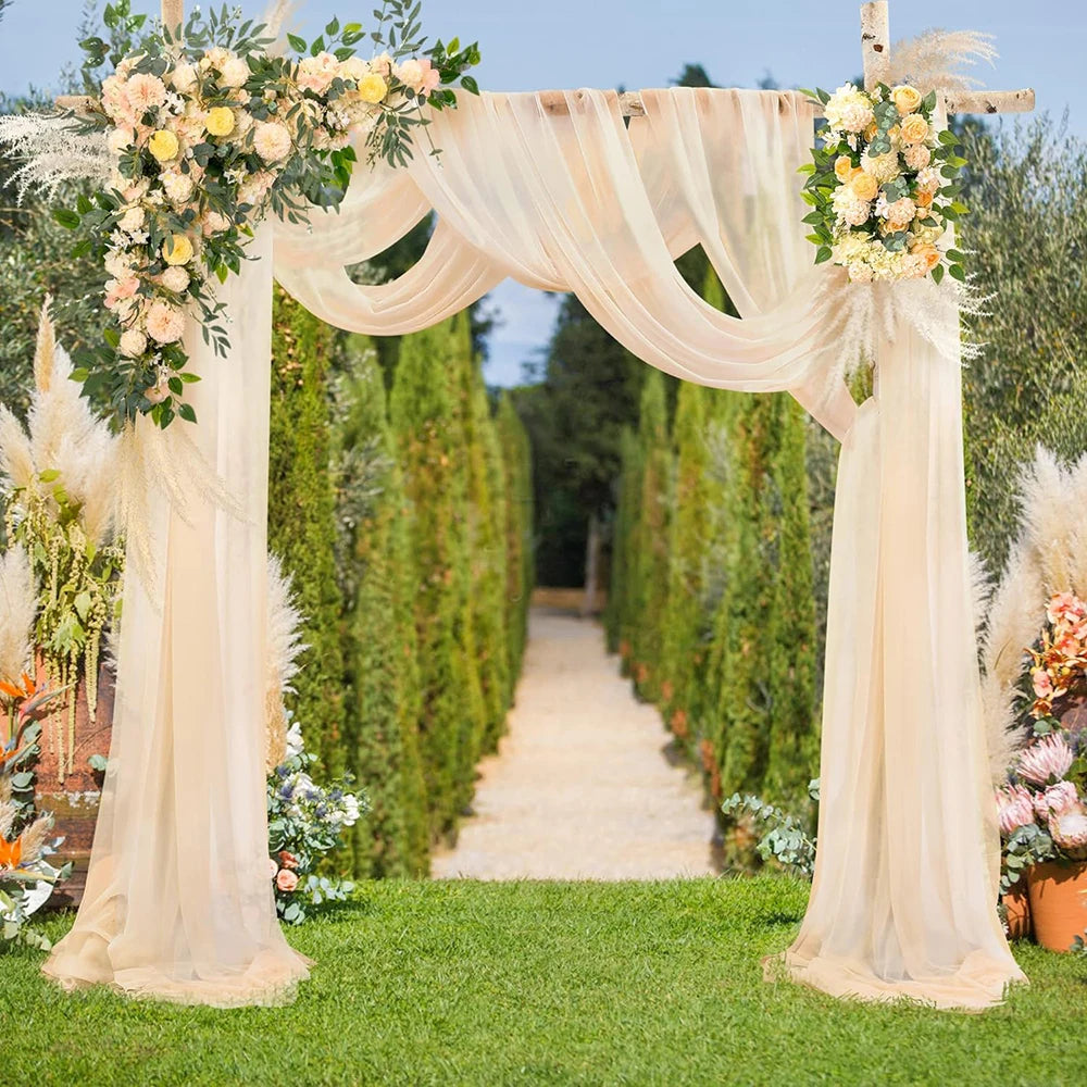 A wedding arch decorated with sheer beige chiffon draping fabric panels and floral greenery in a garden setting.