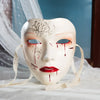A white masquerade mask with red blood-like markings and decorative patterns, displayed on a white cloth.