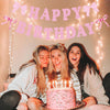 Three young women smiling while holding a glitter pink bow birthday banner in front of them.